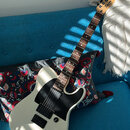 Bílá elektrická kytara s černým pickguardem a hnědým hmatníkem. Na hlavě je viditelné logo Squier a nápis STRATOCASTER. Kytara má dva humbuckery, jed…