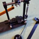 Černý kovový stojan s mechanismem pro napnutí strun, který obsahuje pružinu a nastavovací šrouby. Vedle stojanu leží samostatná černá kroužková součá…