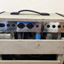 Zadní strana kytarového zesilovače v šedém krytu s černou rukojetí. Na kovovém panelu jsou viditelné nápisy MAINS IN, 220/240V 1.2A, 120V 1.3A a MAIN…