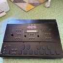 Černý sampler Ensoniq ASR-X Advanced Sampler/Resampler. Na horním panelu je vidět různé ovládací prvky, tlačítka a displej, přičemž uprostřed je nale…