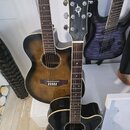Tři kytary opřené o bílou stěnu. V popředí je černá akustická kytara s bílým lemováním a logem YAMAHA na hlavě. Za ní je hnědá akustická kytara s pře…