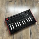 Černý kompaktní MIDI kontrolér s červeným rámem, umístěný na dřevěné podlaze. Na horní části je nápis AKAI PROFESSIONAL a v dolní části model MPKmini…