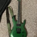 Zelená elektrická kytara s kovovými strunami a dvěma černými humbuckery. Na hlavě je viditelné logo Ibanez. Kytara má zelený popruh a vedle ní leží č…