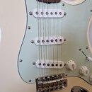 Bílá elektrická kytara s bledě zeleným pickguardem a hnědým hmatníkem. Nástroj má tři bílé snímače typu single-coil a tři bílé ovládací knoflíky s ná…