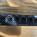Černý rackový přístroj s řadou konektorů XLR. Nad konektory jsou bílé nápisy OUTPUT 16, OUTPUT 15, OUTPUT 14, OUTPUT 13, OUTPUT 12 a OUTPUT 11. Na sp…