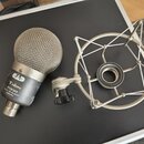 Dynamický mikrofon s kovovou síťkovou hlavou a šedým tělem. Na těle je nápis TRION 7000 DUAL ELEMENT PERSON a logo ve tvaru stylizovaného písmena T.…