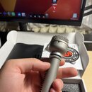 Ruka drží stříbrný mikrofon s kovovou síťkou. Na prstenci pod hlavou mikrofonu je nápis "SM58". V pozadí je vidět bílá krabice a štítek s červeným lo…