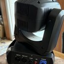 Černý světelný efekt typu moving head z plastu a kovu. Na zadní straně jsou konektory s nápisy DMX IN, DMX OUT a POWER, dále držák pojistky s nápisem…