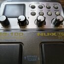 Stříbrný kytarový procesor s displejem a ovládacími knoflíky. Na přístroji je žlutý nápis NUX MG-100 a text TRUE ANALOG. Pod knoflíky jsou popisky SE…