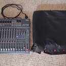 Šedý mixážní pult s nápisy YAMAHA a MX12/4. Zařízení obsahuje několik faderů, knoflíků a vstupních konektorů. Vedle mixážního pultu je černý přepravn…