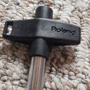 Černý plastový klíč v tvaru kříže s kovovým hrotem. Na plastové části je vyražen nápis Roland. Předmět leží na světlém koberci.