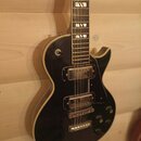 Černá elektrická kytara s tvarem single-cutaway a bílým lemováním kolem těla. Nástroj má dva humbuckery, čtyři ovládací knoflíky a přepínač snímačů.…