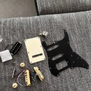 Sada komponentů pro elektrickou kytaru rozložená na šedém textilním povrchu. Sada obsahuje černý pickguard, krémovou zadní krytku, dva krémové snímač…