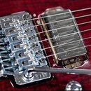 Detail elektrické kytary s červeným lakem a viditelnou strukturou dřeva. Na snímku je chromovaný kobylník s tremolem a dvěma humbuckery s kovovými kr…