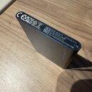 Fotografie zachycuje zadní stranu externího pevného disku v tmavém provedení. Na povrchu jsou bílé nápisy: MODEL MU-PC1T0T, SN S6XDN5OWA1D608M, RATED…