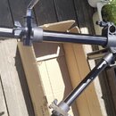 Černý kovový stojan na bicí nástroje s teleskopickými rameny a utahovacími šrouby. Nástroj leží na dřevěném podkladu vedle otevřeného kartónového box…