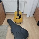 Akustická kytara s přírodním dřevem, světlým topem a tmavým hnedým rándolem. Na hlavě je viditelné logo Martin. Kytara stojí na černém stojanu a vedl…