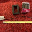 Na červeném koberci je položen stříbrný humbucker s černými cívkami a připojeným kabelem. Vedle něj je černá pěnová vložka a krabička s nápisem Seymo…