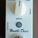 Bílý kytarový efekt s nápisem Hustle Drive a značkou MOUER. Na horní straně jsou dva černé knoflíky s popisky VOLUME a TONE, uprostřed velký bílý kno…