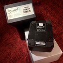 Na fotografii je černý efekt pedál s logem Dream 65 REVERB-AMP, který leží na bílém kartonu. Vedle něj je černá krabice s nápisem Dream 65 REVERB-AMP…
