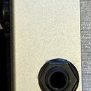 Detail stříbrného kovového šasi efektního pedálu. Jsou vidět tři konektory: jeden malý napájecí vstup a dva větší zásuvky pro jack kabel s černými pl…