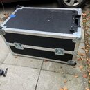 Černý transportní kufr s hliníkovými hranami a rohovými zpevněními. Má čtyři kolečka, dvě kovové zámky na přední straně a jednu černou plastovou ruko…