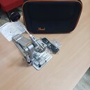 Stříbrný bicí pedál s černým šlapákem a přiloženým manuálem. Na černé přepravní tašce s oranžovým lemováním je viditelné logo Pearl. Vedle pedálu je…