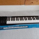 Černý MIDI kontrolér s klaviaturní klávesou, položený na modré krabici. Na přístroji je v horní části bílý nápis ALESIS a v pravém rohu označení V49.…