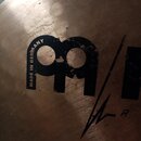 Detailní záběr na povrch kovového činely v bronzové barvě. Je zde vidět černý nápis MADE IN GERMANY a část velkého loga tvořeného dvěma písmeny. V pr…