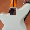 Zadní strana elektrické kytary v bílé barvě s hnědým krčkem. Na kovovém spojovacím plátku mezi tělem a krčkem je vyryto logo Squier. Povrch laku vypa…