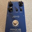 Modrý kytarový efekt s nápisy JOYO a ARGOS OVERDRIVE. Na horní straně jsou čtyři černé ovládací knoflíky s popisky ATTACK, TONE, GATE a LEVEL. Pod ni…