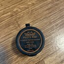 Kulatá černá plastová nádobka s oranžovým potiskem. Na víčku je logo koruny a nápisy REMO, PUTTY PAD, To Remove, Squeeze Container, NON-TOXIC, REMO,…