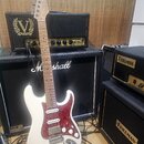 Bílá elektrická kytara s červeným vzorovaným pickguardem, umístěná před dvěma zesilovači. Na hlavě kytary je viditelné logo Fender. V pozadí jsou zes…
