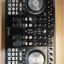 Černý DJ 컨트롤러 s názvem TRAKTOR KONTROL S4. Zařízení obsahuje dva velké stříbrné jog-wheels, faderové posuvníky a množství ovládacích tlačítek a knofl…