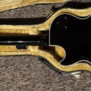 Černá kytara s gold hardwarem v otevřeném hnědém plišovém kufru. Na zadní straně těla je zlatý nápis Epiphone Gibson. Na hmatníku je vidět malý černý…
