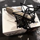 Stříbrný kondenzátorový mikrofon v černém pavoukovém držáku, položený na bílém originálním pude. Na těle mikrofonu je nápis RODE a na krabici jsou vi…