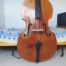 Dřevěný smyčkový nástroj, pravděpodobně kontrabas, v hnědé barvě. Povrch vykazuje známky opotřebení a drobné škrábance, zejména v dolní části těla. N…