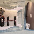 Bílá pickguard s instalovanou elektronikou a třemi černými snímači, umístěná v průhledném plastovém obalu. Na jedné z komponent je viditelné logo EMG…