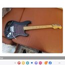 Černá elektrická kytara s tmavě fialovým vzorovaným pickguardem a světlou hnedou hmatníkovou deskou. Na hlavě kytary je viditelné logo Fender. Kytara…