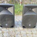 Dvě černé akustické reproboxy z plastu s hornovými výstupníky v horní části. Na přední mřížce každého reproduktoru je umístěno bílé kulaté logo s pse…