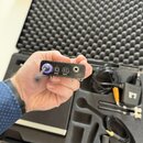 Ruka drží malý černý elektronický přístroj s ovládacími prvky. Na přední straně jsou vidět fialový knoflík, nápisy SYNC a MUTE u tlačítek a jeden kon…
