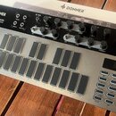 Stříbrný analogový syntetizátor s černým ovládacím panelem. Na horní části je nápis DONNER SYN BASS ANALOG SYNTHESIZER AND SEQUENCER a logo DONNER. P…