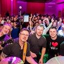 Fotografie zachycuje skupinu lidí v sále, v popředí jsou čtyři muži. První muž zleva drží elektrickou kytaru s bílým hmatníkem a logem Gibson na hlav…