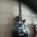 Černá elektrická kytara s tvarem single-cutaway a bílým lemováním kolem korpusu. Nástroj má dva humbuckery, Tune-o-matic můstek a čtyři ovládací knof…