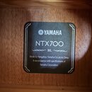 Detailní záběr na čtvercový černý štítek s roundedrohy vlepený do dřevěného těla nástroje. Štítek obsahuje bílé nápisy: logo YAMAHA, model NTX700 a o…