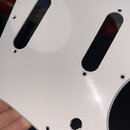 Bílá plechovka (pickguard) na kytaru s dvěma oválnými výřezy pro snímače. Kolem otvorů jsou viditelné montážní dírky na šrouby. V dolní části je kula…