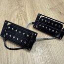 Dvě černé humbuckery s montážními rámečky, položené na dřevěném povrchu. Snímače mají viditelné kovové šroubky a pólové piny. Na fotce nejsou žádné č…
