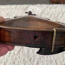 Detailní pohled na hranu spodní desky smyčcového nástroje z tmavého dřeva. Na hraně je viditelný kulatý otvor a kovový držák na rameno. Povrch dřeva…