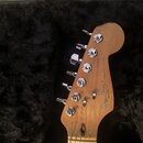 Hlava elektrické kytary z přírodního dřeva s chromovanými ladičkami, položená v černém plišovém kufru. Na dřevě je vidět bílý nápis Fender STRATOCAST…