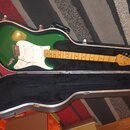 Zelená elektrická kytara v černém tvrdém kufru. Na hlavě hředu je viditelné logo Fender. Kytara má bílý pickguard a světle hnědý javorový hmatník. Na…